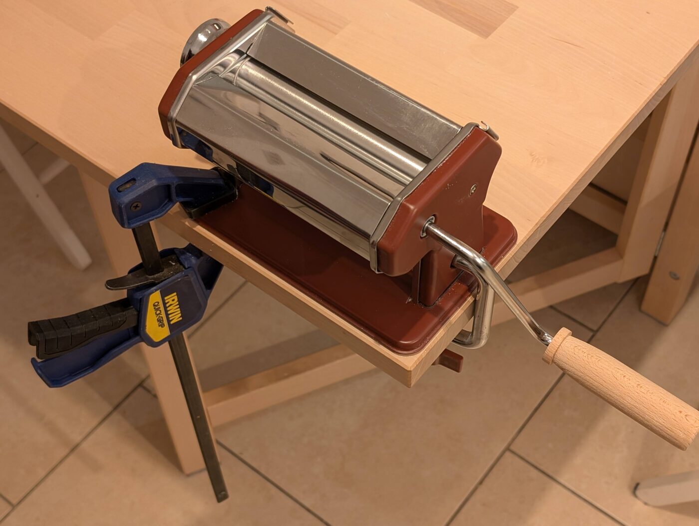 A hand-cranked pasta machine clamped to the edge of a wooden table with a blue Irwin Quick-Grip bar clamp