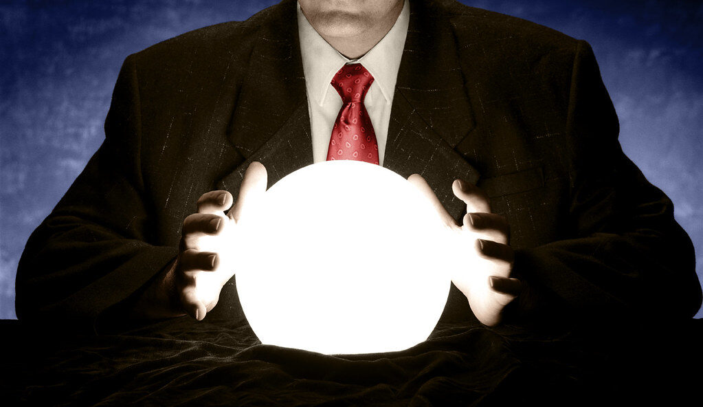 A businessman in a suit and red tie, hands cupped around a brightly glowing crystal ball on a velvet-draped table. Photo: InfoWire.dk, Flickr, public domain