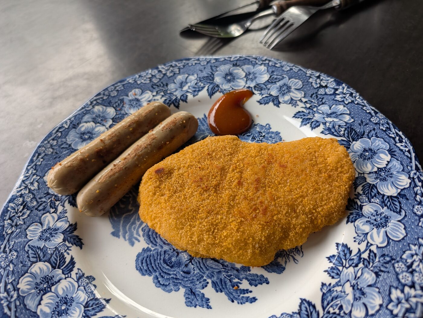 A breaded vegan schnitzel and two vegan sausages on a plate, with a dollop of brown sauce